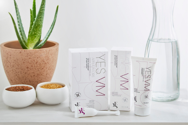 YES VM vaginal moisturiser range on a white background and white surface. An aloe vera plant and a pitcher of water are positioned behind. Two small bowls of flax seed are next to the tube of YES VM and the applicator.