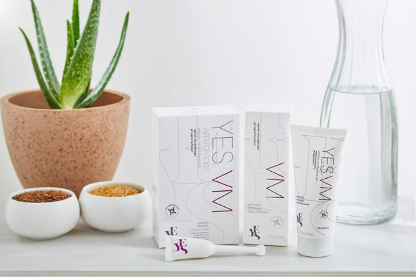 YES VM vaginal moisturiser range on a white background and white surface.  An aloe vera plant and a pitcher of water are positioned behind.  Two small bowls of flax seed are next to the tube of YES VM and the applicator.  